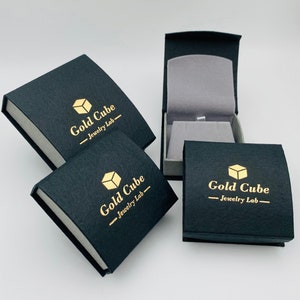 May include: Four black jewelry boxes with gold lettering and a gold cube logo. One box is open, revealing a gray interior. The boxes are arranged on a white surface. The text on the boxes reads "Gold Cube Jewelry Lab".