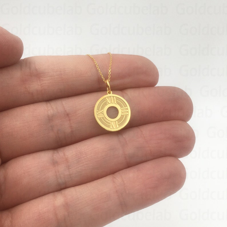 May include: A gold-colored pendant necklace featuring a circular charm with Roman numerals and a central design. The charm is suspended from a delicate gold chain. The necklace is displayed against a neutral background.