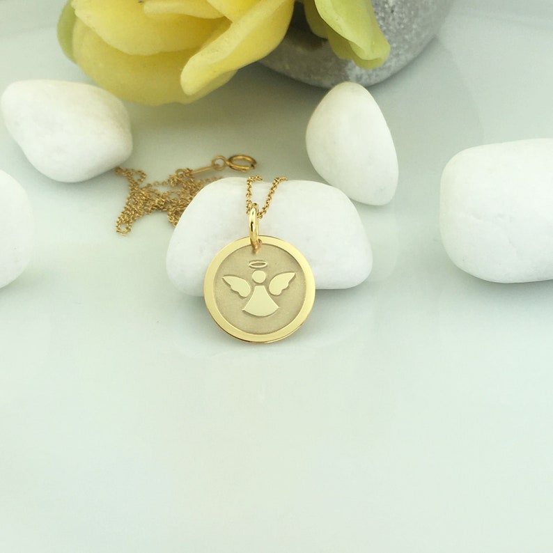 May include: A gold-colored pendant necklace featuring a circular charm with an angel design. The angel has outstretched wings and a halo. The necklace chain is also gold-colored, and the pendant is resting on a white stone.