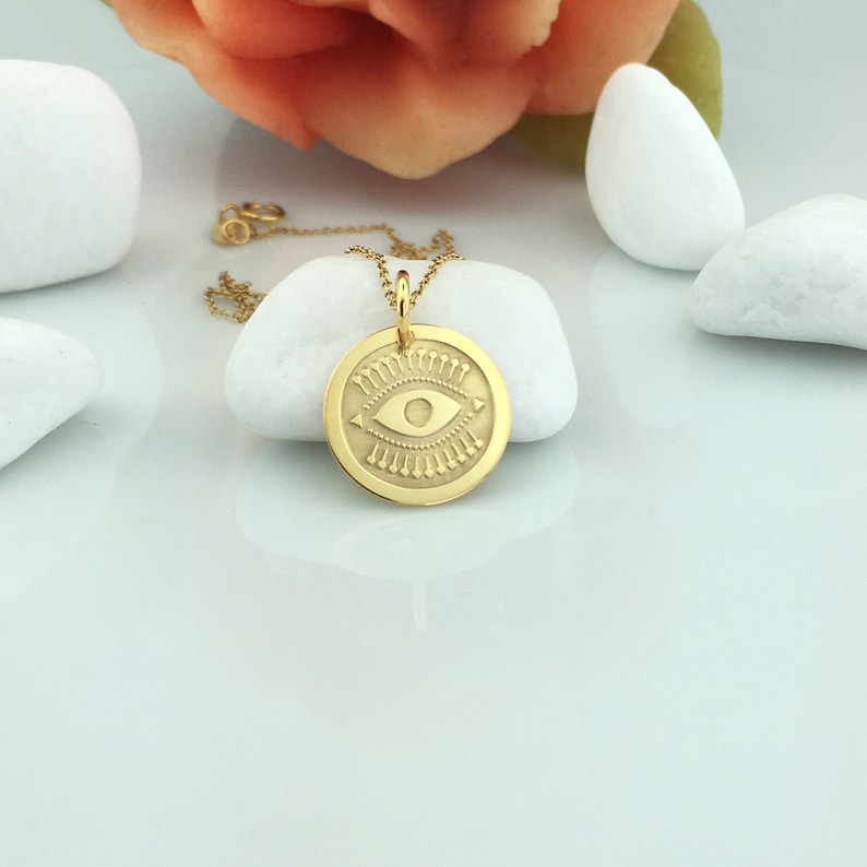 Pode incluir: Um colar com pingente dourado com um berloque circular com um design de olho. O berloque tem um olho detalhado no centro, rodeado por elementos decorativos. O colar &eacute; exibido em uma superf&iacute;cie branca com pedras decorativas e uma flor ao fundo.