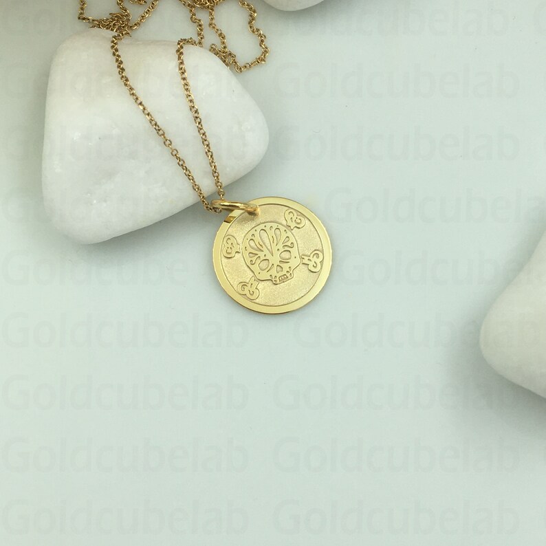 May include: A gold chain necklace with a round pendant featuring a detailed sugar skull design. The pendant is on a white background.