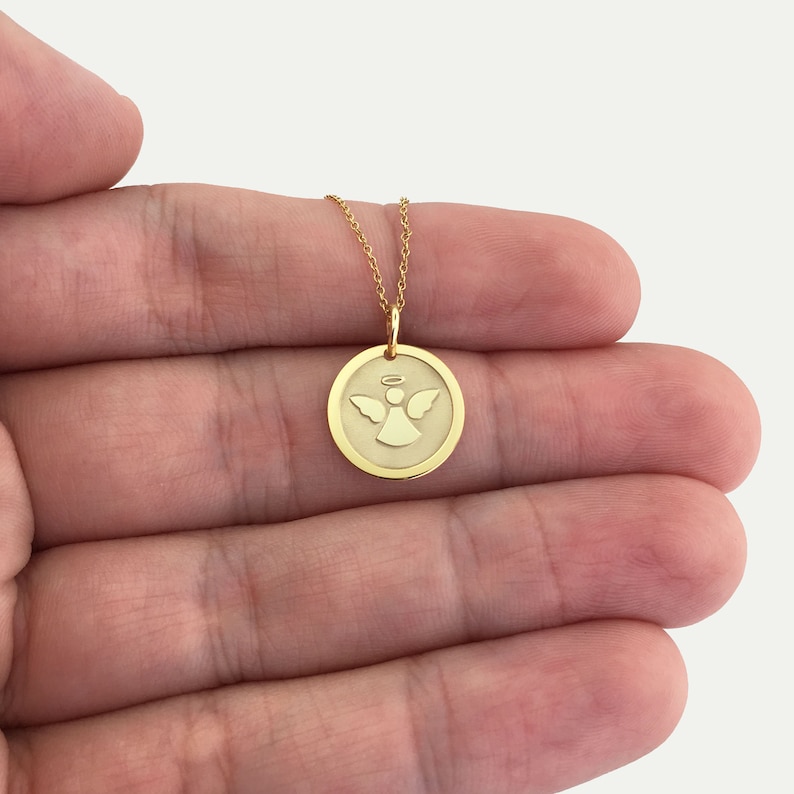 May include: A gold-colored necklace featuring a circular pendant with an engraved angel design. The angel has outstretched wings and a halo. The pendant hangs from a delicate gold chain. The necklace is displayed against a light background.