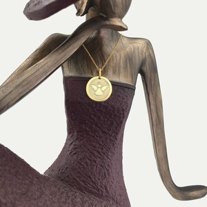 May include: A gold-colored pendant necklace featuring a circular charm with an angel design. The necklace is displayed on a bronze-colored sculpture of a woman wearing a dark purple dress. The pendant has a halo above the angel.