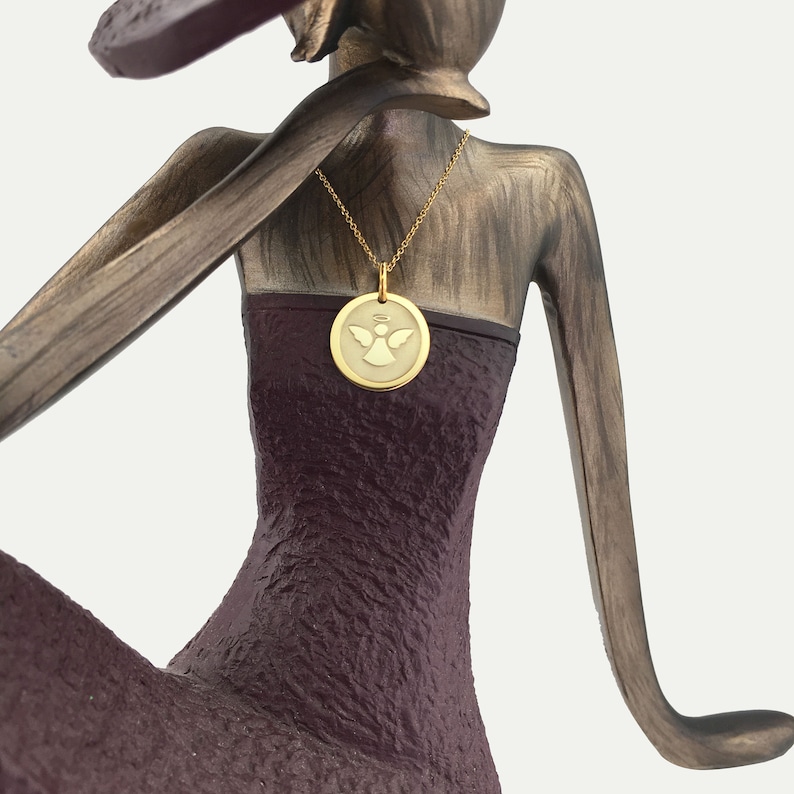 May include: A gold-colored pendant necklace featuring a circular charm with an angel design. The necklace is displayed on a bronze-colored sculpture of a woman wearing a dark purple dress. The pendant has a halo above the angel.