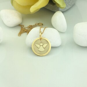 May include: A gold-colored pendant necklace featuring a circular charm with an angel design. The angel has outstretched wings and a halo. The necklace is displayed on a white stone, with a gold chain and clasp.