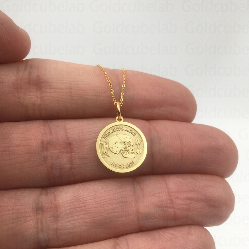 K&ouml;nnte beinhalten: Goldfarbene Halskette mit einem runden Anh&auml;nger. Der Anh&auml;nger zeigt ein Totenkopf- und Stundenglas-Design mit den Gravuren "MEMENTO MORI" und "AMOR FATI". Die Kette wird vor einem hellen Hintergrund pr&auml;sentiert.