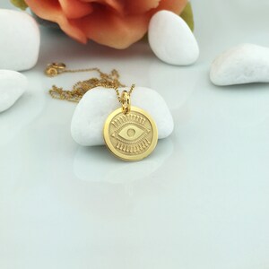 Pode incluir: Um colar com pingente dourado com um berloque circular gravado com um desenho de olho. O pingente pende de uma corrente delicada. O colar &eacute; exibido em uma superf&iacute;cie branca com pedras brancas e uma flor ao fundo.