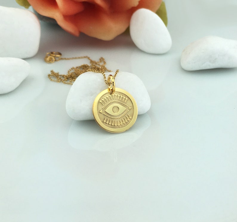 Pode incluir: Um colar com pingente dourado com um berloque circular gravado com um desenho de olho. O pingente pende de uma corrente delicada. O colar &eacute; exibido em uma superf&iacute;cie branca com pedras brancas e uma flor ao fundo.