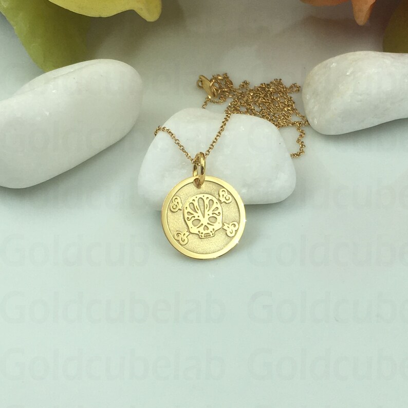 May include: A gold chain necklace with a round pendant featuring a detailed skull design. The pendant has a crossbones pattern around the edge.