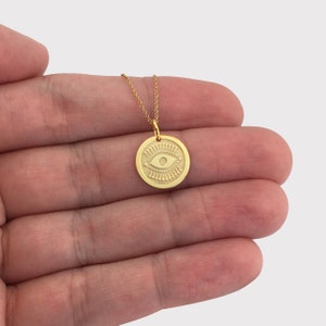 Pode incluir: Colar dourado com um pendente redondo com um design de olho gravado. A corrente &eacute; delicada. A joia &eacute; exibida em um fundo neutro.