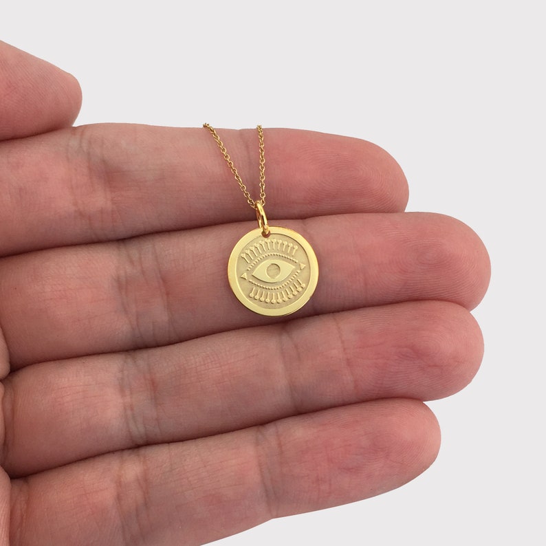 Pode incluir: Colar dourado com um pendente redondo com um design de olho gravado. A corrente &eacute; delicada. A joia &eacute; exibida em um fundo neutro.