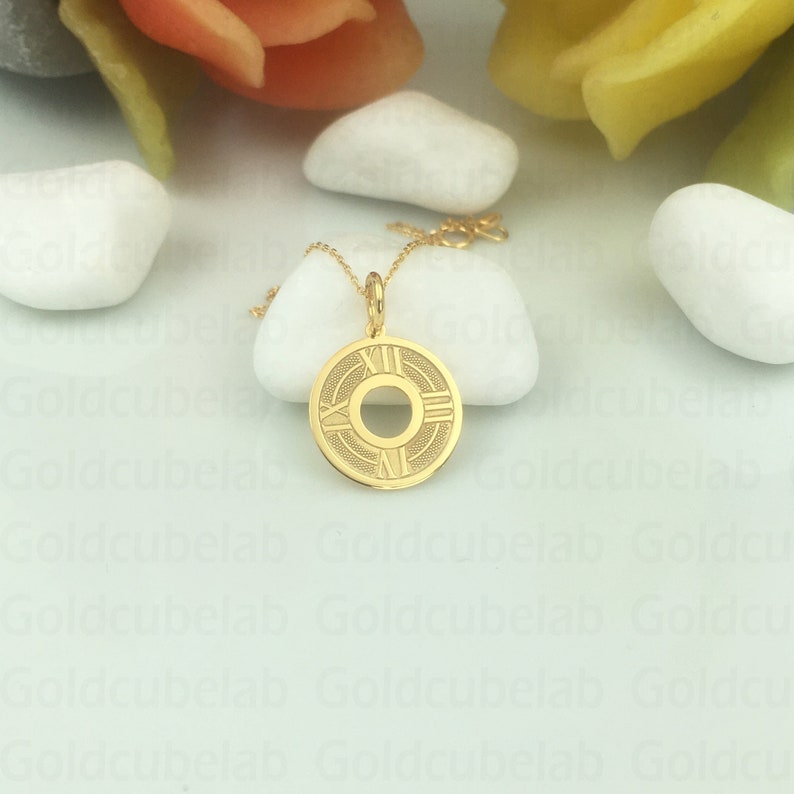 May include: A gold-colored pendant necklace featuring a circular pendant with Roman numerals and a central design. The pendant hangs from a delicate gold chain. The necklace is displayed on a white surface.