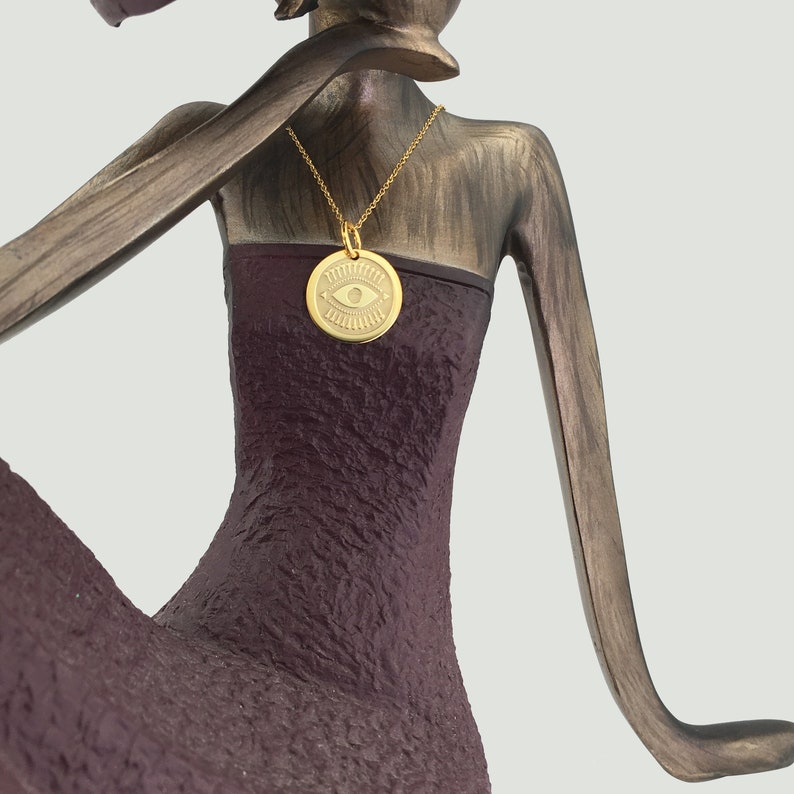 Pode incluir: Um colar com pingente dourado com um berloque circular com um design de olho. O colar pende de uma corrente dourada sobre um vestido roxo escuro. A est&aacute;tua tem um acabamento bronze.