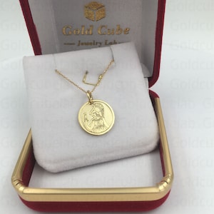 May include: A gold-colored pendant necklace featuring a circular charm with an engraved image of a figure in a helmet. The necklace is displayed in a red jewelry box with a white interior. The box has gold trim and the text "Gold Cube Jewelry Lab".