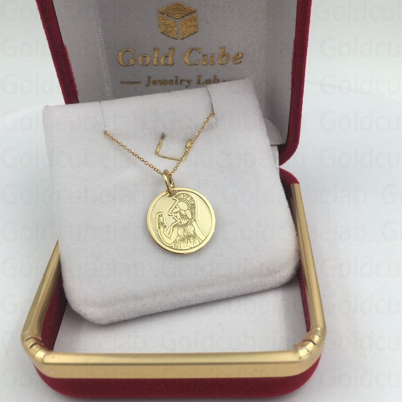May include: A gold-colored pendant necklace featuring a circular charm with an engraved image of a figure in a helmet. The necklace is displayed in a red jewelry box with a white interior. The box has gold trim and the text "Gold Cube Jewelry Lab".