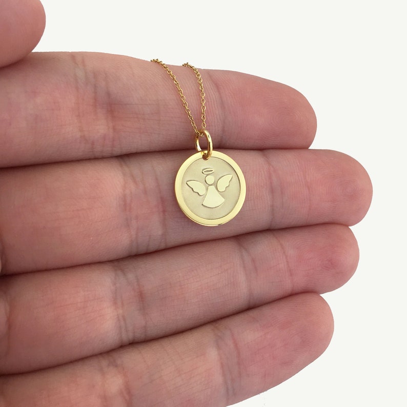 May include: A gold-colored pendant necklace featuring a circular charm with an engraved angel design. The angel has wings and a halo. The necklace has a delicate chain.