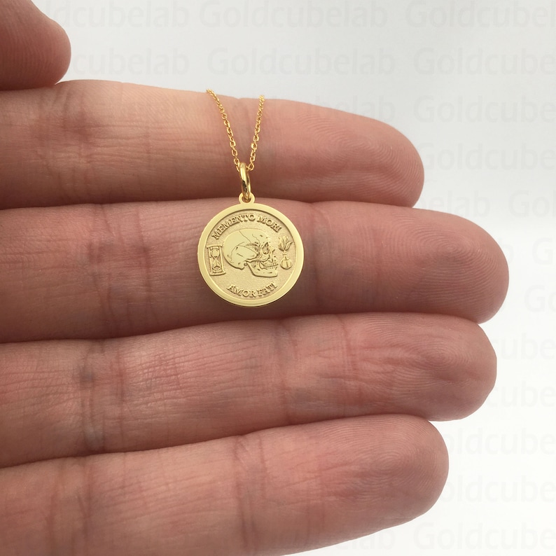 K&ouml;nnte beinhalten: Eine goldfarbene Halskette mit einem Anh&auml;nger. Der runde Anh&auml;nger zeigt einen Totenkopf, die Worte "Memento Mori" und "Amor Fati", eine Sanduhr und weitere kleine Details. Die Kette ist filigran. Der Anh&auml;nger ist goldfarben.