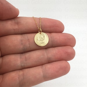 May include: A gold-colored pendant necklace featuring a circular charm with an engraved image of a figure in a helmet. The charm is suspended from a delicate gold chain. The necklace is displayed against a neutral background.