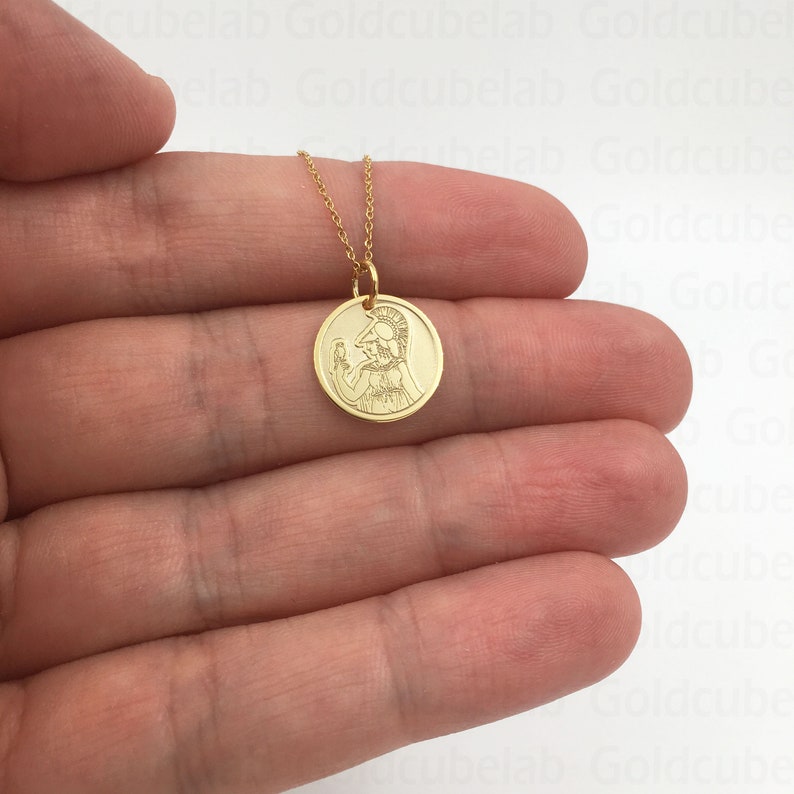 May include: A gold-colored pendant necklace featuring a circular charm with an engraved image of a figure in a helmet. The charm is suspended from a delicate gold chain. The necklace is displayed against a neutral background.