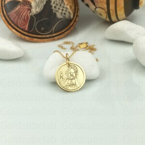 May include: A gold-colored pendant necklace featuring a circular charm with a detailed engraved image of a figure in classical attire. The pendant hangs on a delicate gold chain, set against a white background.