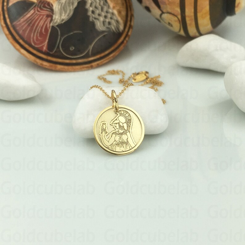 May include: A gold-colored pendant necklace featuring a circular charm with a detailed engraved image of a figure in classical attire. The pendant hangs on a delicate gold chain, set against a white background.
