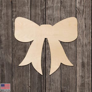 May include: A large, unfinished wooden bow cutout, perfect for crafting and DIY projects. The bow is light-colored and has a classic bow shape with two loops and two long tails. The background is a rustic wooden surface.