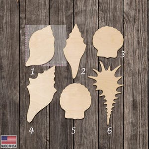 May include: Six wooden seashell cutouts in various shapes and sizes, arranged on a weathered wood surface. The cutouts are unpainted and feature natural wood grain. The image includes the text "MADE IN USA" with the American flag.