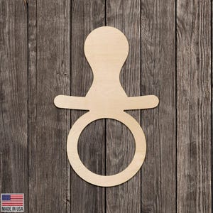 Puede incluir: Un recorte de madera con forma de chupete, con una parte superior redondeada, una barra horizontal y una base circular. El chupete es de madera de color claro y está sobre un fondo de madera rústica. Se ven las palabras "MADE IN USA".