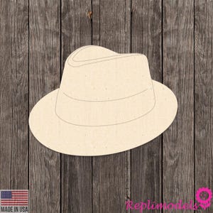 Könnte beinhalten: Ausgeschnittener Fedora-Hut in hellem Beige, mit breiter Krempe und strukturierter Krone. Der Hut besteht aus einem hellfarbigen Material und ist vor einem dunklen Holzmaserungshintergrund platziert. Die Worte "MADE IN USA" sind sichtbar.