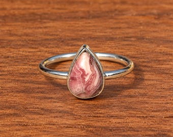 Puede incluir: Un anillo de plata con una piedra preciosa de rodocrosita en forma de lágrima. La piedra muestra un patrón jaspeado de remolinos rosas y blancos. La banda de metal es fina, pulida y circular, sujetando la piedra en un engaste sencillo.