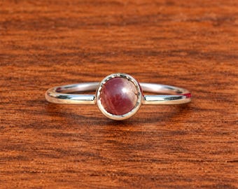Puede incluir: Un anillo de plata con una piedra preciosa de color rojo oscuro, redonda y tallada en cabujón, engastada en un bisel liso. La banda es fina, pulida y metálica, mostrando un diseño sencillo que resalta el color carmesí de la piedra central.
