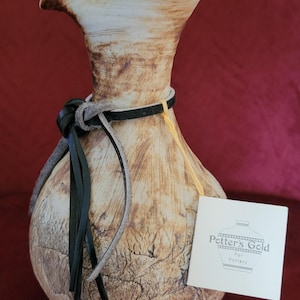 May include: A light brown ceramic vase with a wide mouth and a narrow neck. The vase has a leather strap around the neck with black tassels. The vase is decorated with a crackled glaze and has a label that reads "Potter's Gold Pottery".