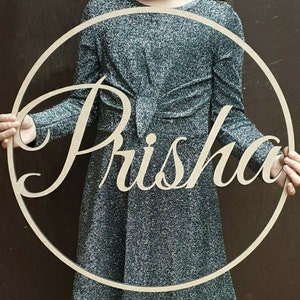 Pu&ograve; includere: Un cerchio di legno con il nome "Prisha" tagliato in un font corsivo. Il cerchio &egrave; tenuto da una persona che indossa un abito argentato scintillante.