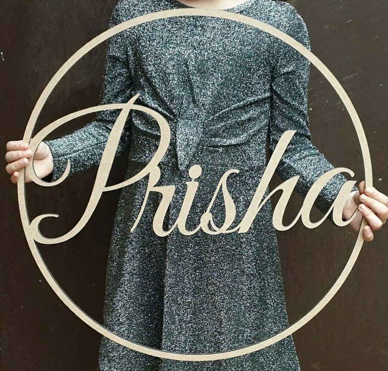 Pu&ograve; includere: Un cerchio di legno con il nome "Prisha" tagliato in un font corsivo. Il cerchio &egrave; tenuto da una persona che indossa un abito argentato scintillante.