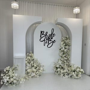 May include: White backdrop with two arches, decorated with white flowers and greenery. A black sign with the text "Bride to Be" is centered on the backdrop.