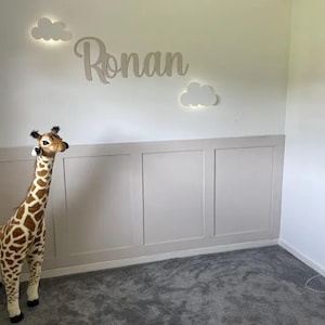 May include: A child's bedroom with a giraffe soft toy. The wall is decorated with the name "Ronan" in cursive lettering and two cloud-shaped lights. The lower wall is painted in a neutral tone, and the floor is covered with grey carpet.