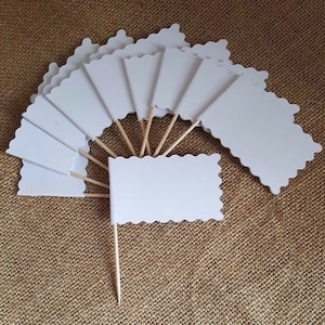 May include: A set of white scalloped flags on wooden sticks. The flags are blank and can be used for a variety of purposes, such as decorating cakes or cupcakes, or as place cards.