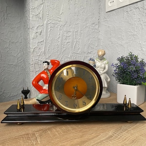 Puede incluir: Un reloj de madera vintage con una esfera dorada y agujas negras. El reloj está sobre una base de madera negra con detalles dorados. El reloj está rodeado de figuritas decorativas.