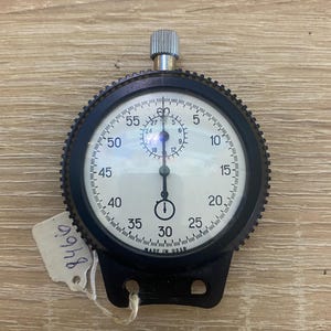 May include: A vintage stopwatch with a black bezel and a white face, featuring black numerals and markings. The stopwatch has a silver winding crown and a black base with two holes. A tag with the number "8466" is attached. The text "MADE IN USSR" is visible.