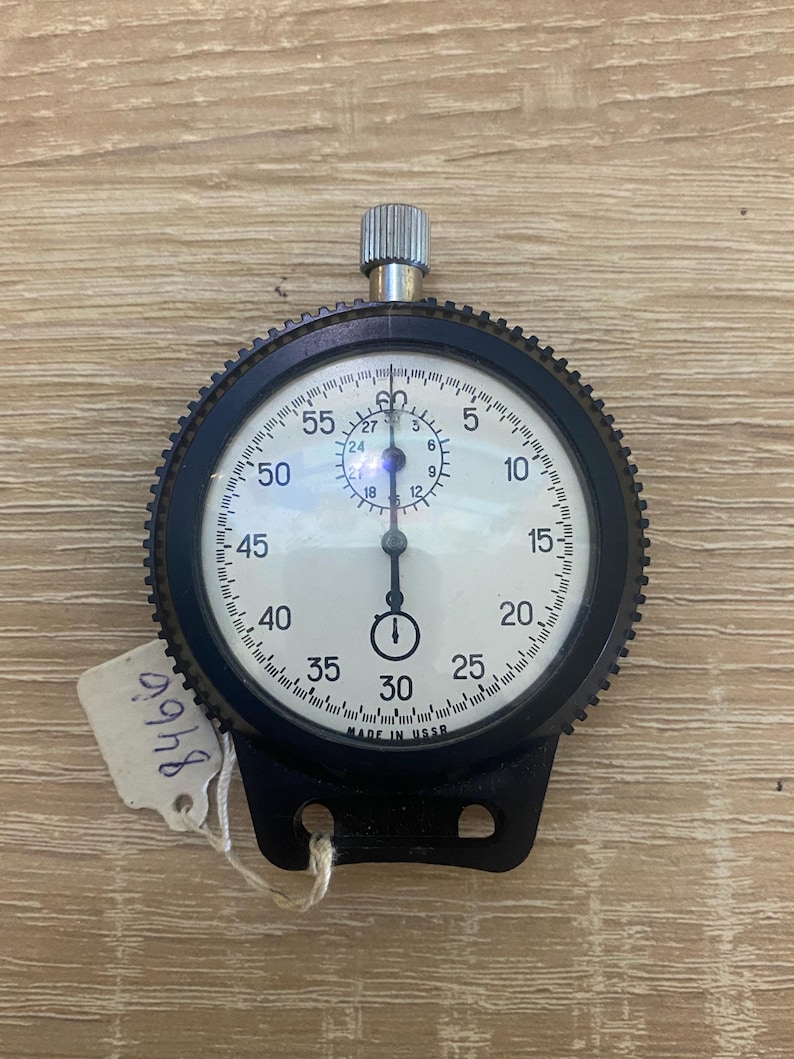 Pu&ograve; includere: Un cronometro vintage con lunetta nera e quadrante bianco, con numeri e segni neri. Il cronometro ha una corona di carica argentata e una base nera con due fori. Un'etichetta con il numero "8466" &egrave; attaccata. Il testo "MADE IN USSR" &egrave; visibile.