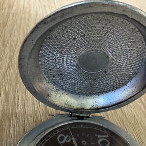 Puede incluir: Un reloj de bolsillo antiguo abierto, con una caja de color plateado. La esfera del reloj es visible, mostrando los n&uacute;meros 8 y 10. El interior de la tapa del reloj tiene un patr&oacute;n texturizado detallado.