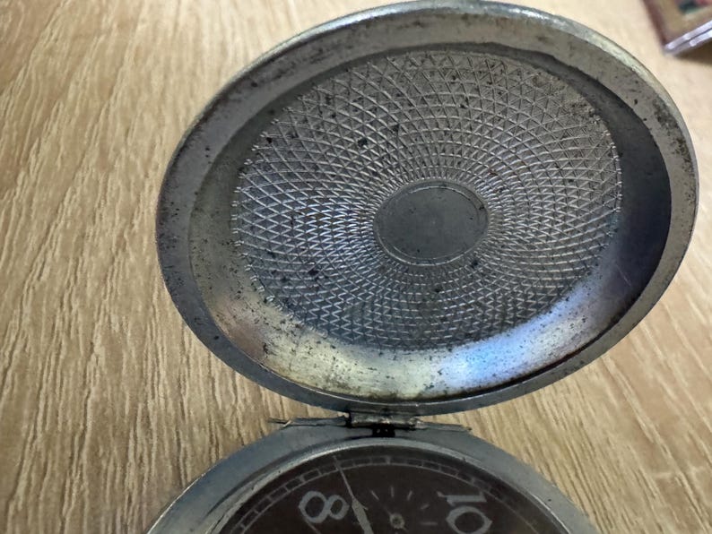 Puede incluir: Un reloj de bolsillo antiguo abierto, con una caja de color plateado. La esfera del reloj es visible, mostrando los n&uacute;meros 8 y 10. El interior de la tapa del reloj tiene un patr&oacute;n texturizado detallado.