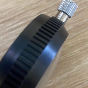 Pu&ograve; includere: Primo piano di un comparatore a quadrante nero con una manopola di regolazione argentata e scanalata. Il corpo del quadrante ha un bordo scanalato. Lo sfondo &egrave; una superficie di legno marrone chiaro.