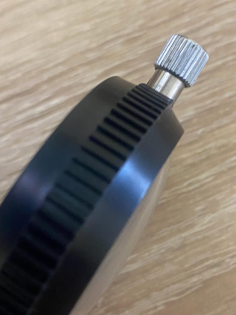 Pu&ograve; includere: Primo piano di un comparatore a quadrante nero con una manopola di regolazione argentata e scanalata. Il corpo del quadrante ha un bordo scanalato. Lo sfondo &egrave; una superficie di legno marrone chiaro.