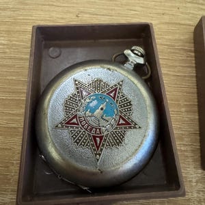 Puede incluir: Reloj de bolsillo plateado vintage con un dise&ntilde;o detallado de estrella en la esfera. La esfera del reloj presenta un emblema rojo y azul con texto cir&iacute;lico. El reloj est&aacute; en una caja rectangular marr&oacute;n.