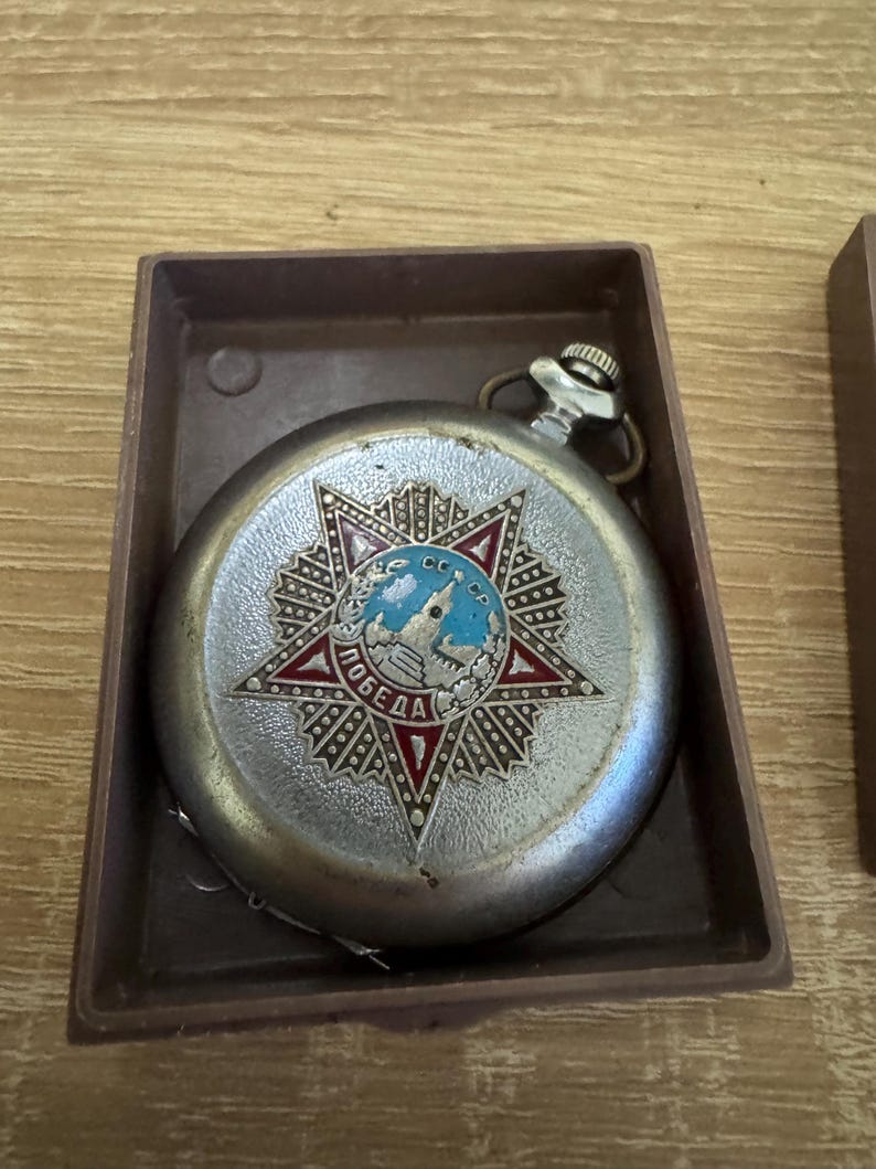 Puede incluir: Reloj de bolsillo plateado vintage con un dise&ntilde;o detallado de estrella en la esfera. La esfera del reloj presenta un emblema rojo y azul con texto cir&iacute;lico. El reloj est&aacute; en una caja rectangular marr&oacute;n.