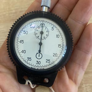 Pu&ograve; includere: Un cronometro vintage con lunetta nera e quadrante bianco con numeri e segni neri. Il cronometro ha una corona argentata e un'impugnatura nera. Il testo "MADE IN USSR" &egrave; visibile sul quadrante.