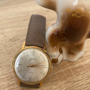 Puede incluir: Un reloj de pulsera dorado con una correa de cuero marrón. La esfera del reloj es blanca con marcas negras y la palabra "СССР" en escritura cirílica.