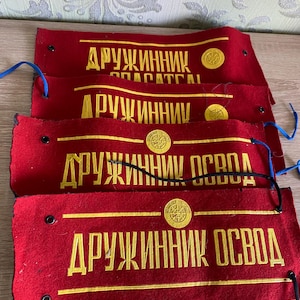 May include: Four red felt armbands with gold embroidered text and a circular emblem. The text reads "ДРУЖИННИК ОСВОД" and other Cyrillic phrases. Each armband has a blue or black string attached.