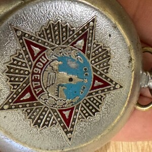 Puede incluir: Reloj de bolsillo plateado con un emblema en forma de estrella que presenta un globo terr&aacute;queo y la palabra "Победа" en letras rojas. El reloj tiene una caja plateada y una tija.